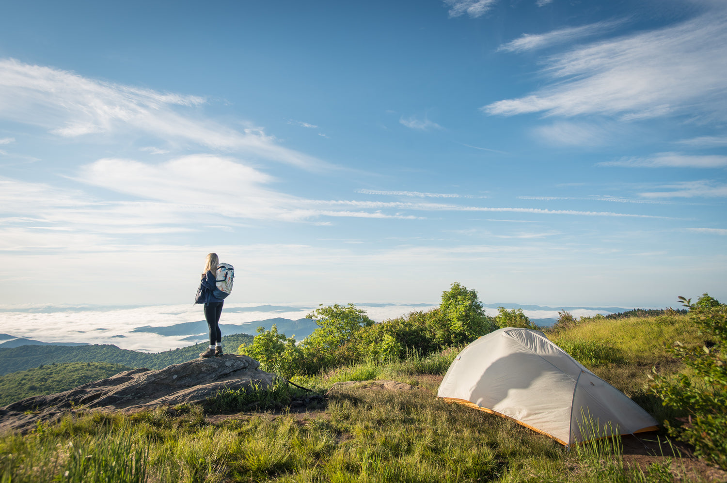 A person with a backpack stands on a rocky outcrop overlooking mountains near a white tent and MYEASYCAMP ultraleicht Isomatte on grassy terrain under a blue sky—perfect for camping adventures.